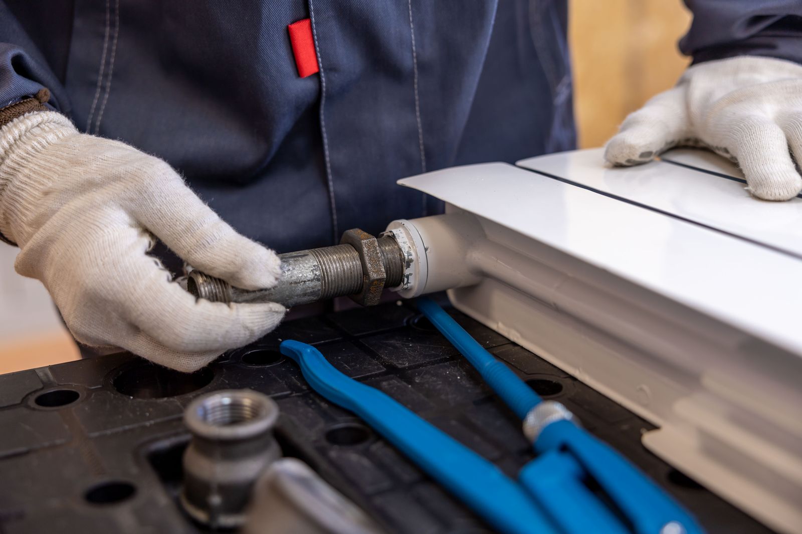 Gloved hands in white cotton work gloves offering a threaded galvanised pipe connector with a hex nut up to a white finned panel, with two blue-handled pipe wrenches and a loose brass fitting laid on a black moulded tray in the foreground.